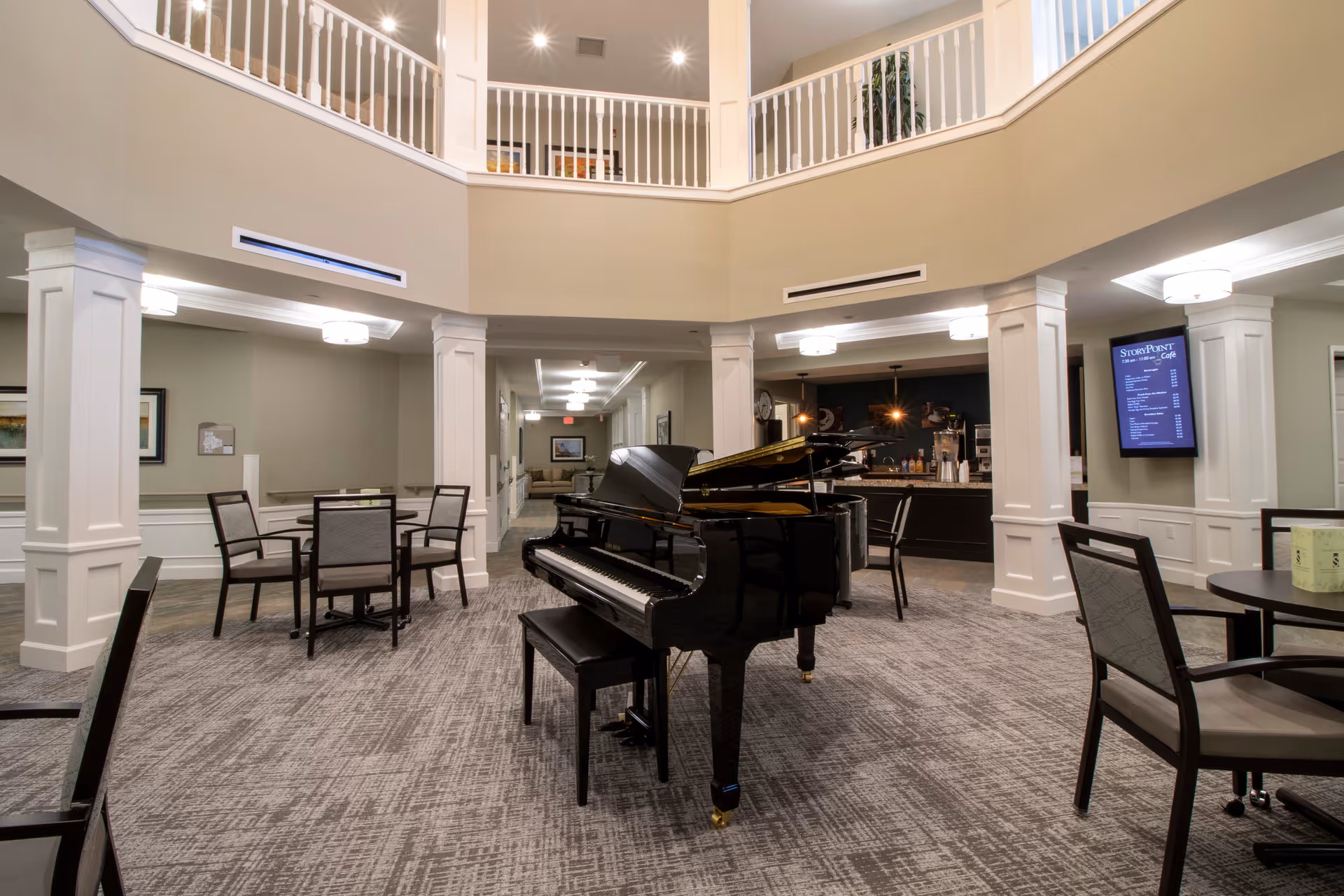 Interior view of a senior living facility common area with a black grand piano in the center, surrounded by tables and chairs. The space features beige walls, white columns, and a carpeted floor. There is a menu board on the wall indicating a café area in the background.