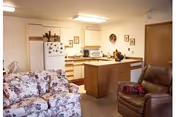 A cozy living area and kitchen in a senior living facility. The living area features a floral-patterned sofa and a brown leather recliner with a red plaid blanket. The kitchen has light wood cabinets, a white refrigerator with magnets, a microwave, and a small island with a decorative centerpiece. The walls are adorned with small framed pictures and a clock.