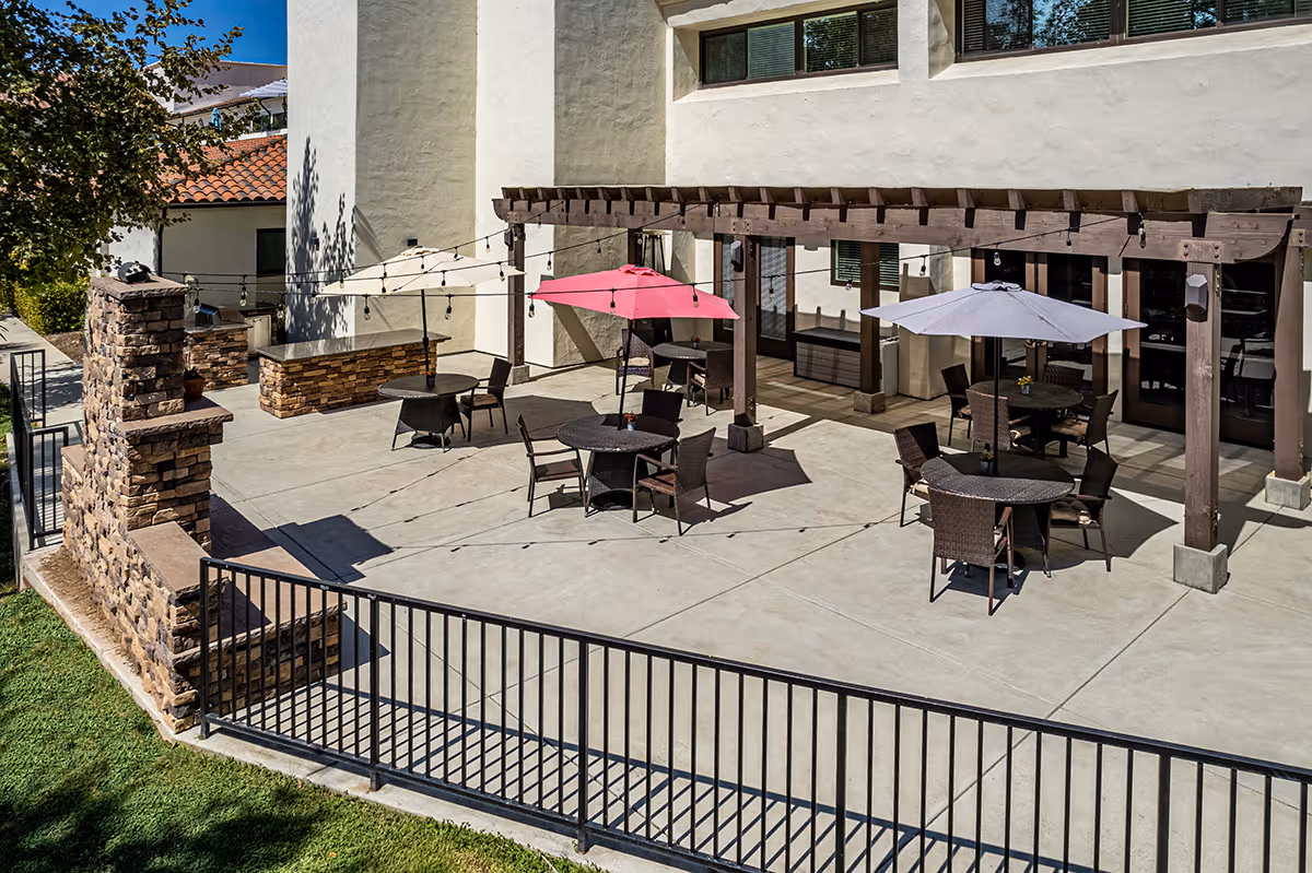 Outdoor patio with round tables, umbrellas, a wooden pergola and string lights outside a senior living building.