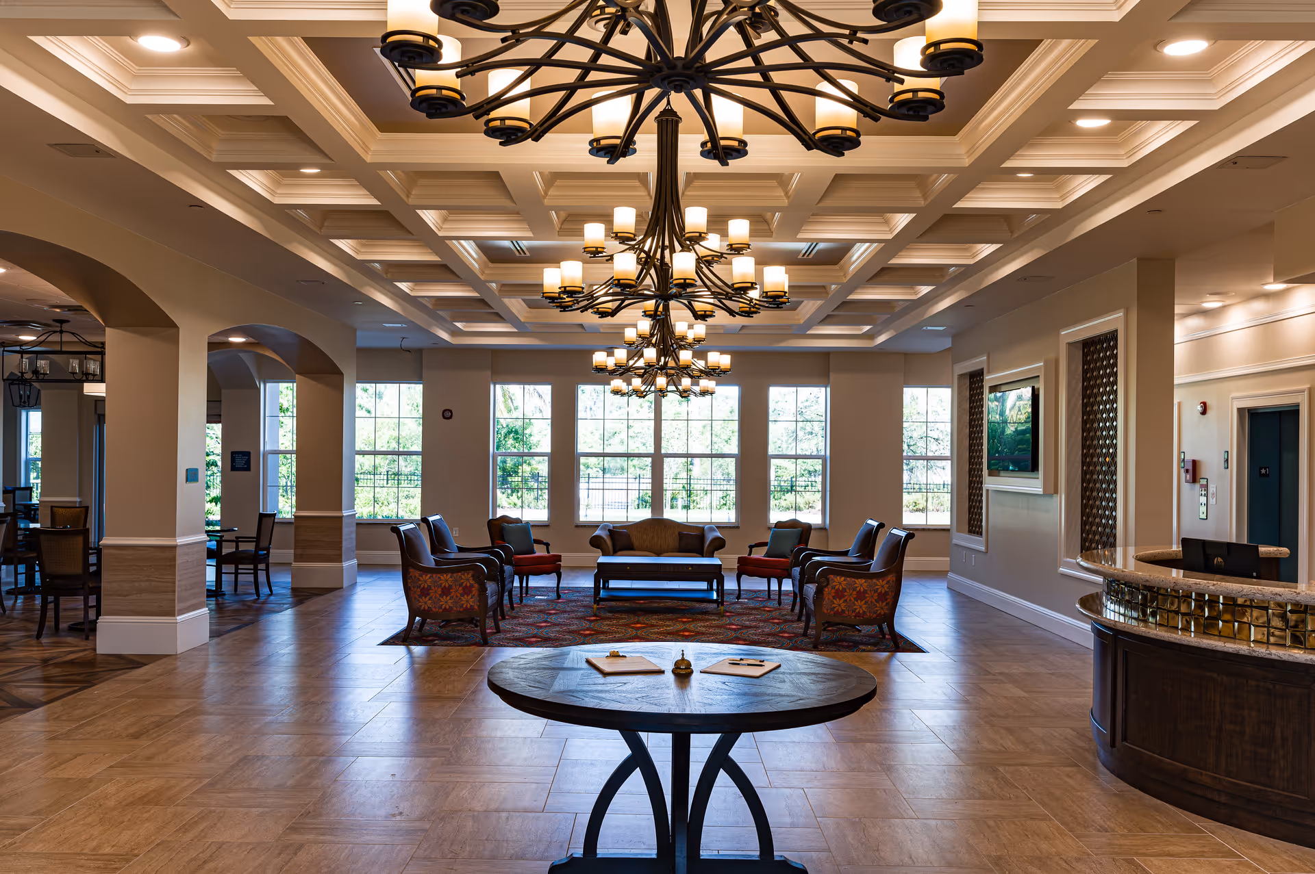 Spacious and elegant senior living facility common area with large windows letting in natural light, multiple chandeliers hanging from a coffered ceiling, a seating area with armchairs and a sofa arranged around a coffee table on a patterned rug, and a round table in the foreground. There is a reception desk to the right and additional seating visible through archways to the left.