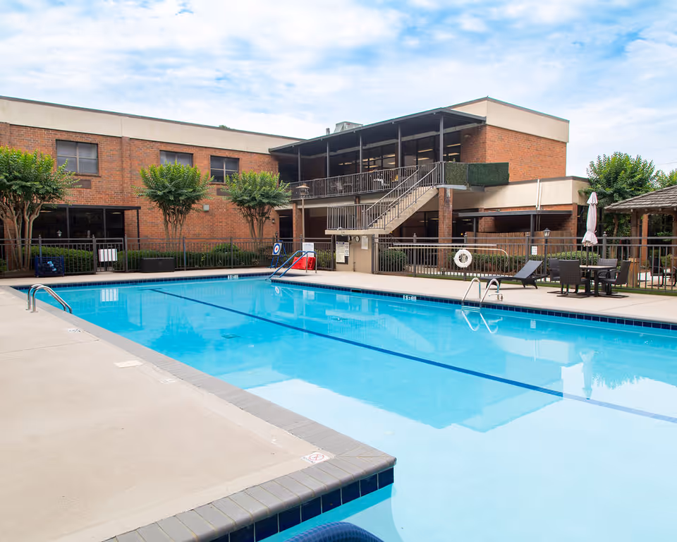 Outdoor swimming pool with clear blue water surrounded by a concrete deck. There are metal handrails for pool entry, lounge chairs, tables with umbrellas, and a two-story brick building with stairs and a balcony in the background. Small trees and shrubs line the perimeter of the pool area.