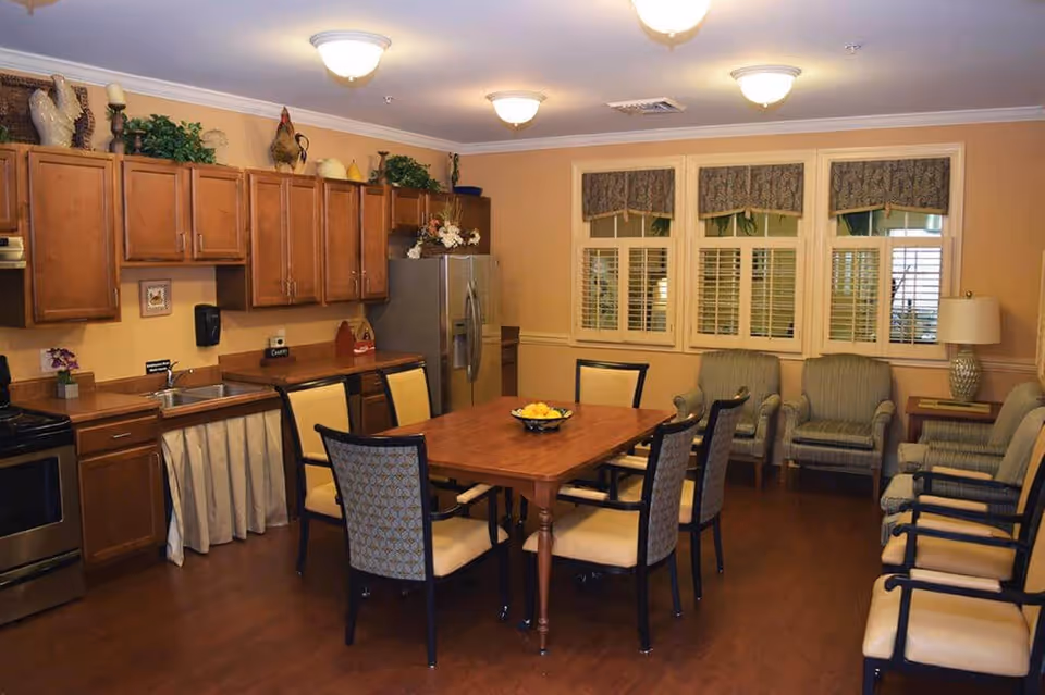 A warm and inviting kitchen and dining area with wooden cabinets, stainless steel refrigerator and stove, a wooden dining table surrounded by six chairs, and additional seating with armchairs along the wall. The room has beige walls, three windows with patterned valances and white shutters, and ceiling lights providing illumination.