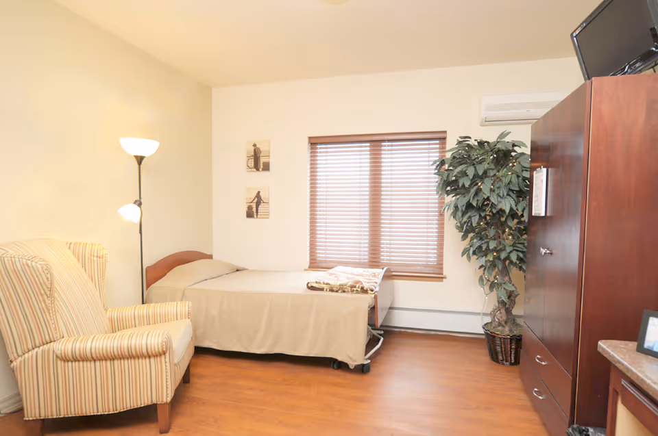 A simple bedroom with a single bed covered in a beige blanket, a striped armchair, a tall floor lamp, two small framed pictures on the wall, a window with wooden blinds, a large potted plant, and a wooden cabinet with a TV on top.