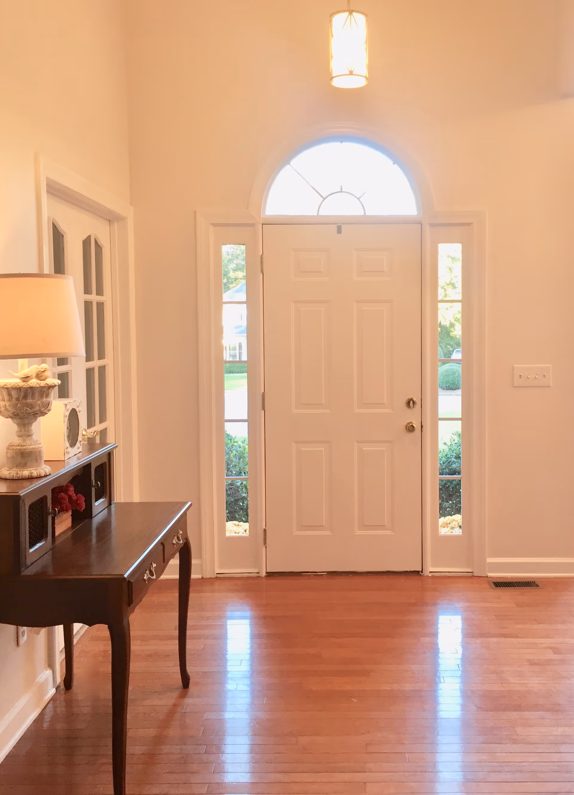 Interior view of an entryway with a white front door featuring sidelights and a semicircular window above. To the left, there is a wooden console table with drawers, a decorative lamp, and a small clock. The floor is hardwood, and the walls are painted light beige.