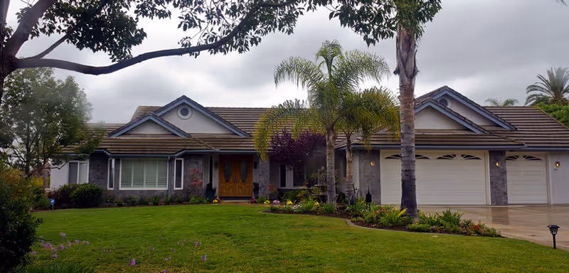 Front exterior of a single-story house with a manicured lawn, palm trees, and a three-car garage under an overcast sky.