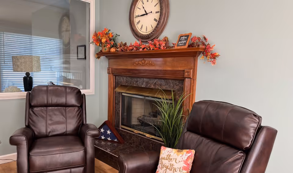 A cozy sitting area with two dark brown leather armchairs facing a wooden fireplace mantel decorated with autumn leaves, small pumpkins, and a round wall clock above it. A green plant is placed in front of the fireplace, and a folded American flag is displayed on the hearth. A window with blinds and a lamp are visible in the background.
