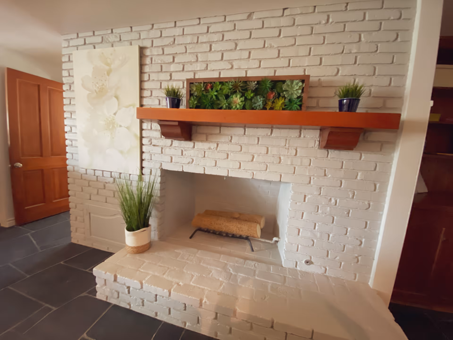 White painted brick fireplace with a wooden mantel holding a rectangular planter with green succulents and two small potted plants. A large floral painting hangs on the left wall above a potted plant on the hearth. A wooden door is visible to the left and dark tiled flooring is seen in front.
