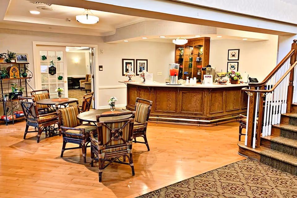 A cozy common area in a senior living facility featuring several round wooden tables with striped cushioned chairs, a wooden reception or refreshment counter with beverage dispensers and cups, decorative plants, framed pictures on the walls, and a carpeted staircase leading upstairs.