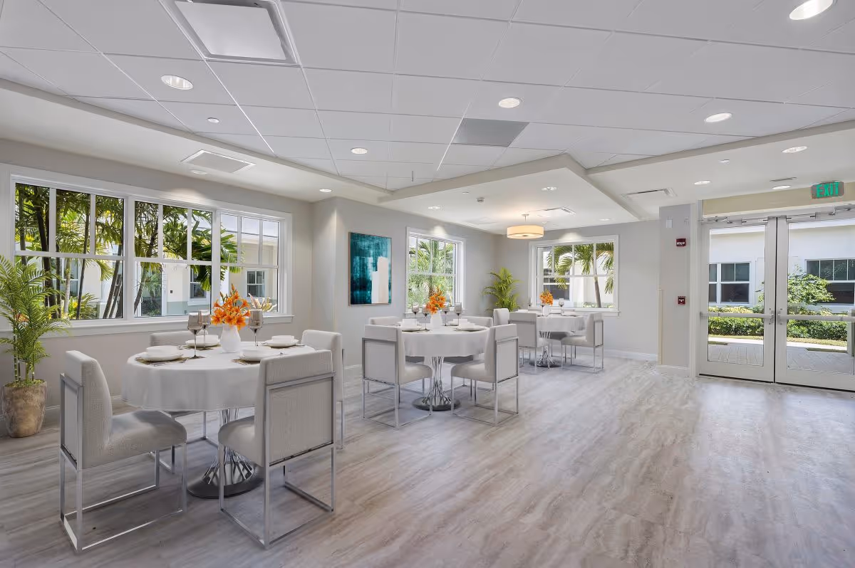 Bright and modern dining room with round tables covered in white tablecloths, each set with plates, glasses, and orange floral centerpieces. The room has large windows letting in natural light and views of greenery outside. Light gray chairs surround the tables, and there are potted plants and a piece of abstract art on the wall. Double glass doors lead outside.