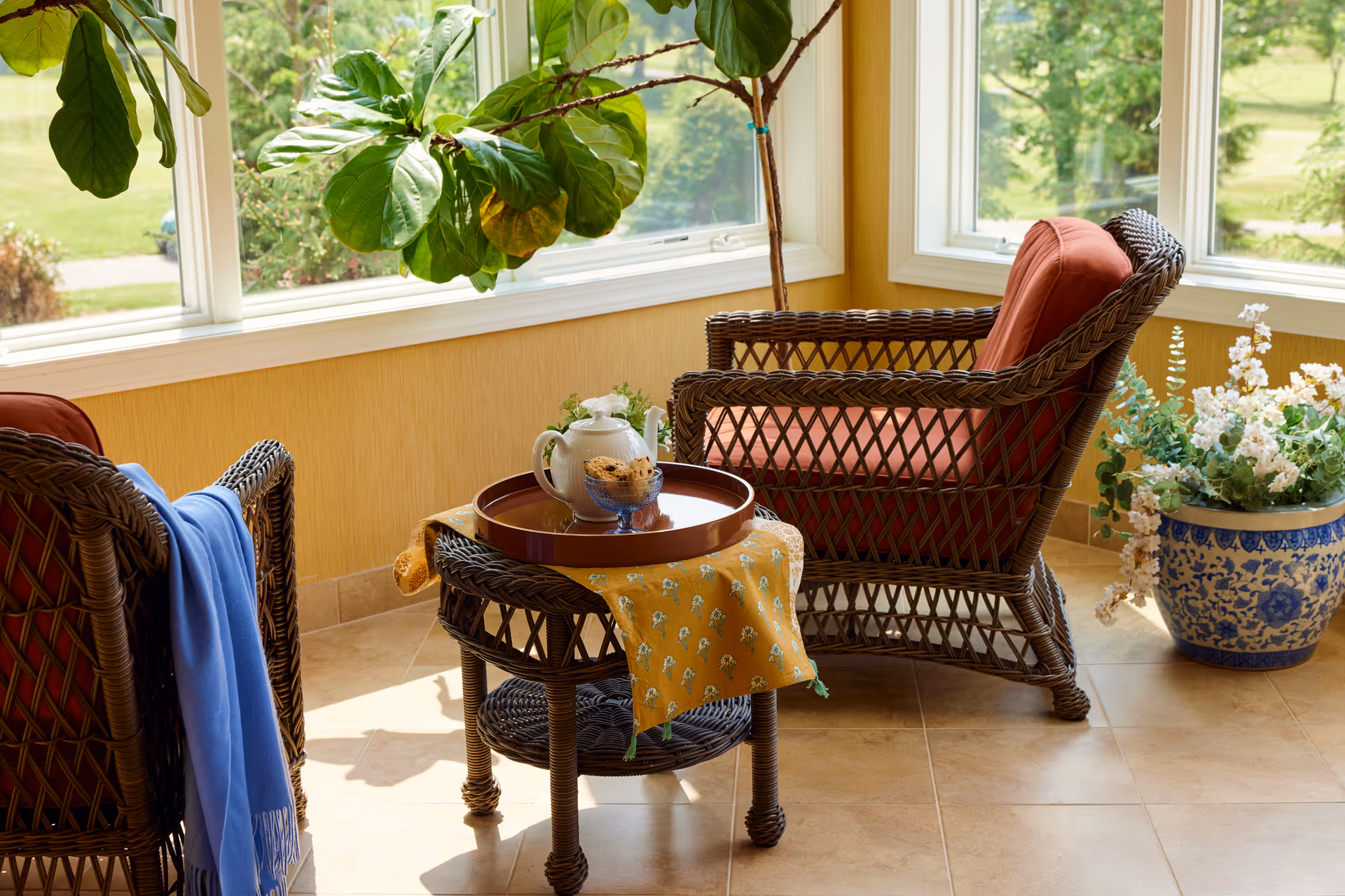 A cozy sunlit sitting area with two wicker chairs featuring red cushions, a small wicker table with a yellow cloth, a tray holding a teapot and a bowl of cookies, a blue throw draped over one chair, large windows showing greenery outside, and a large potted plant with white flowers on the tiled floor.