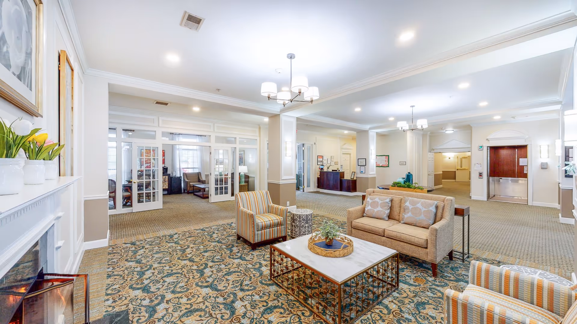 A spacious and well-lit senior living facility common area with beige walls and carpeted floors. The room features a seating area with a beige sofa, two striped armchairs, a white marble coffee table with a decorative tray, and a small round side table. There is a fireplace on the left with white vases and flowers on the mantel. In the background, there are glass-paneled double doors leading to another sitting area with additional chairs and a coffee table. The ceiling has recessed lighting and two modern chandeliers. A reception desk is visible further back, along with an elevator and hallway entrance.