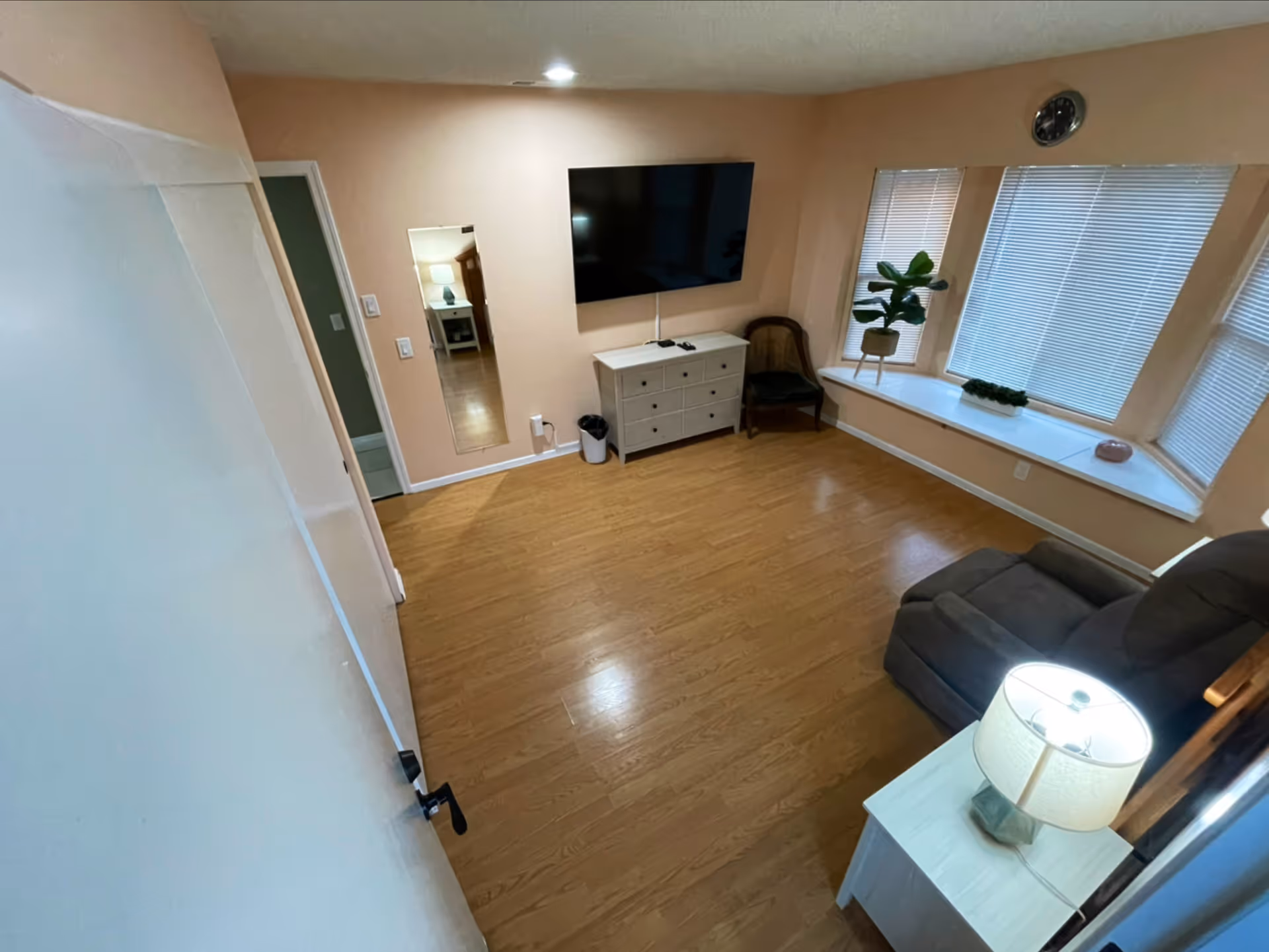 Living room in a senior living facility with a wall-mounted TV, dresser, chair by the window, sofa, and a side-table lamp on a wooden floor.