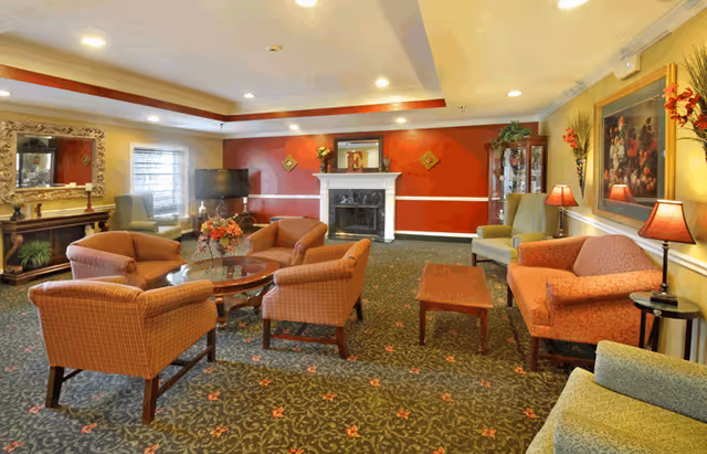 A cozy living room with patterned carpet, several upholstered armchairs and sofas arranged around a glass coffee table, a fireplace with a mantel decorated with a framed letter E and small plants, a large mirror above a wooden console table, a flat-screen TV, and warm lighting from ceiling lights and table lamps.