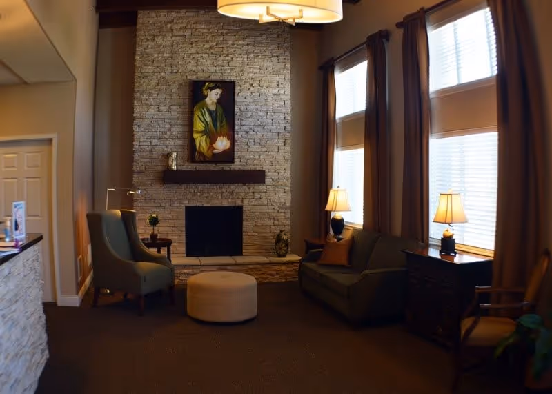 A cozy senior living facility common area with a stone fireplace, a painting above the mantle, two armchairs, a round ottoman, a sofa, two table lamps on side tables, and large windows with brown curtains.