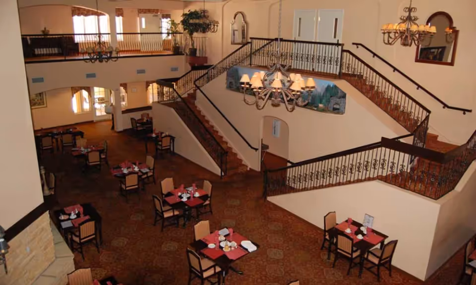 Large interior dining area with multiple set tables, a central chandelier and a grand staircase leading to an upper balcony.