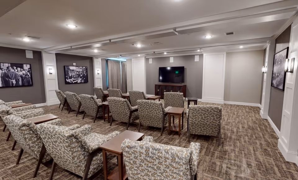 A senior living facility common room with multiple patterned armchairs arranged in rows facing a flat-screen TV mounted on the wall. The room has carpeted floors, neutral-colored walls with white paneling, framed black and white photographs, and soft lighting fixtures on the walls.