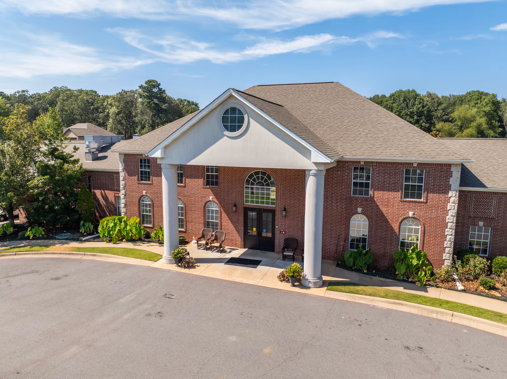 Brick two-story senior living building front with large white entrance columns, arched windows and a circular window under a blue sky.