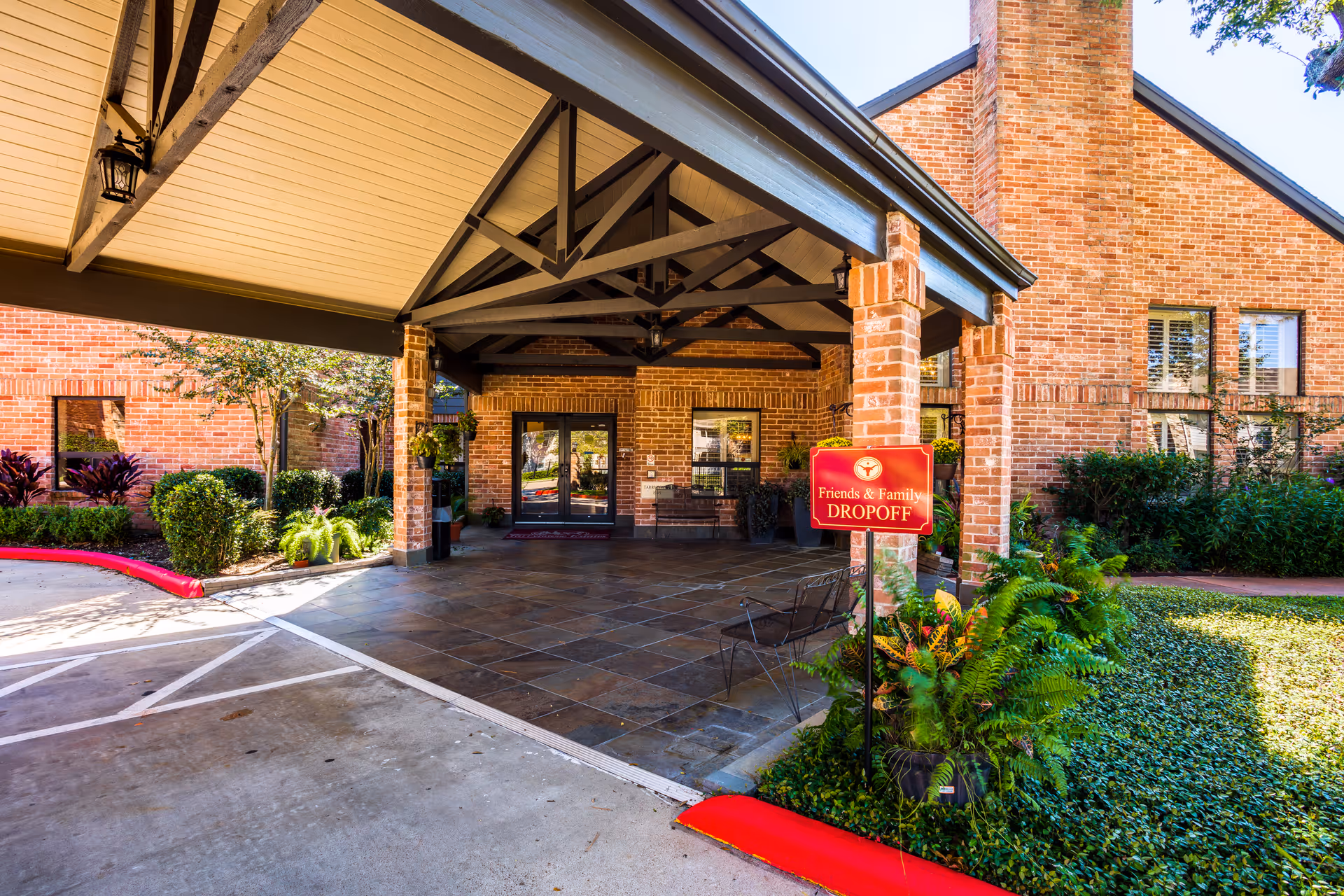 Covered drop-off area at the entrance of a brick building with a sign that reads 'Friends & Family DROPOFF'. The area has a tiled floor, some chairs, and is surrounded by greenery and landscaping.