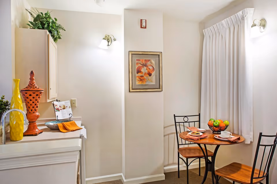 Compact dining nook with a round table set for two beside a small kitchenette counter adorned with decorative vases and a framed floral painting.