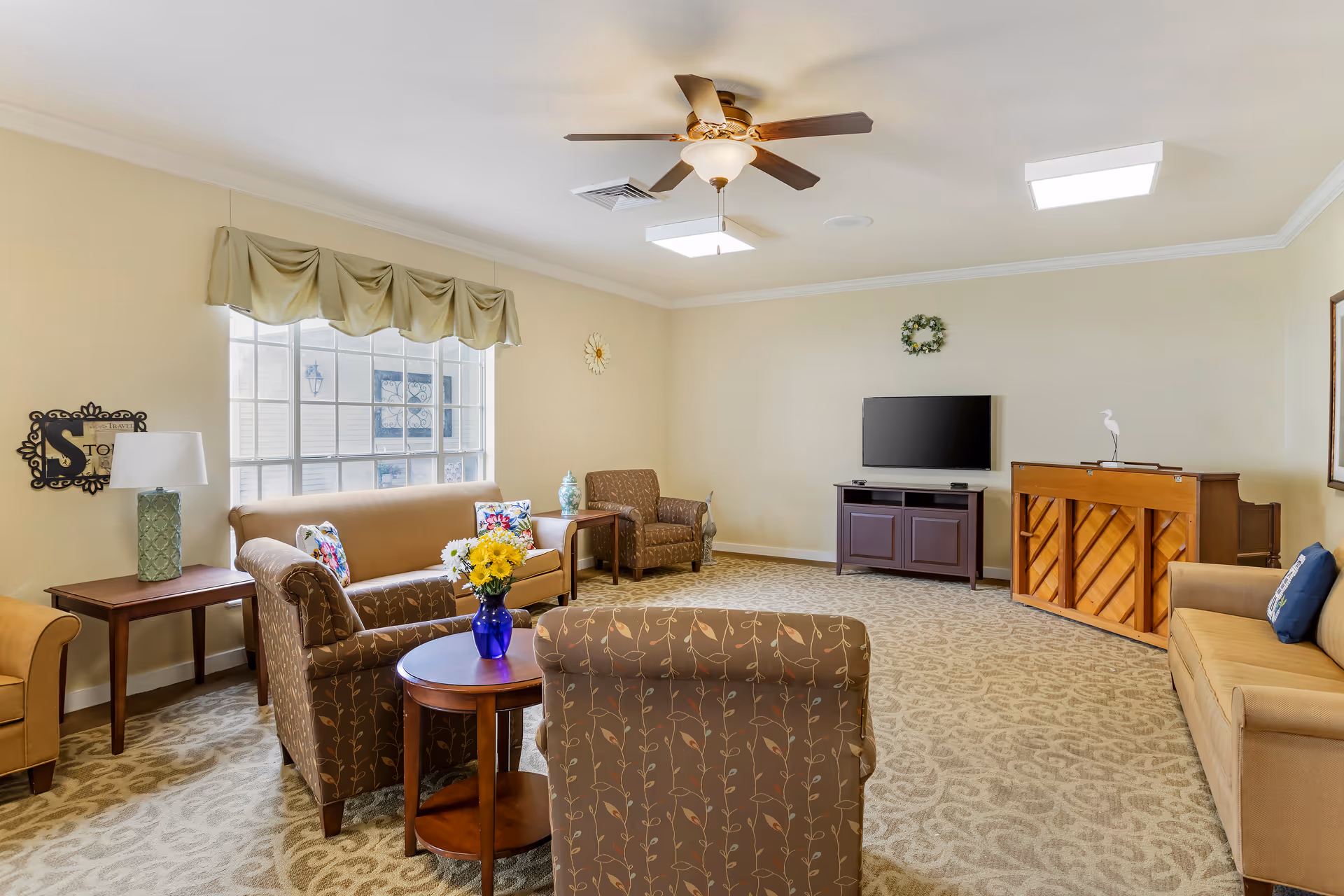 A bright and spacious living room in a senior living facility with beige walls and patterned carpet. The room features multiple upholstered chairs and sofas arranged around wooden side tables, a ceiling fan with light, a flat-screen TV mounted on a dark wooden cabinet, and an upright piano against the far wall. A large window with a beige valance lets in natural light, and decorative items such as a vase with flowers, a small statue, and wall hangings add a homey touch.