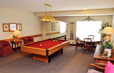A cozy indoor recreational room featuring a red felt pool table in the center, surrounded by comfortable seating including two red armchairs and a wooden table with four chairs near a window. The room is warmly lit with a hanging light fixture above the pool table and table lamps on side tables. Pool cues are mounted on the wall, and decorative framed pictures and plants add to the inviting atmosphere.