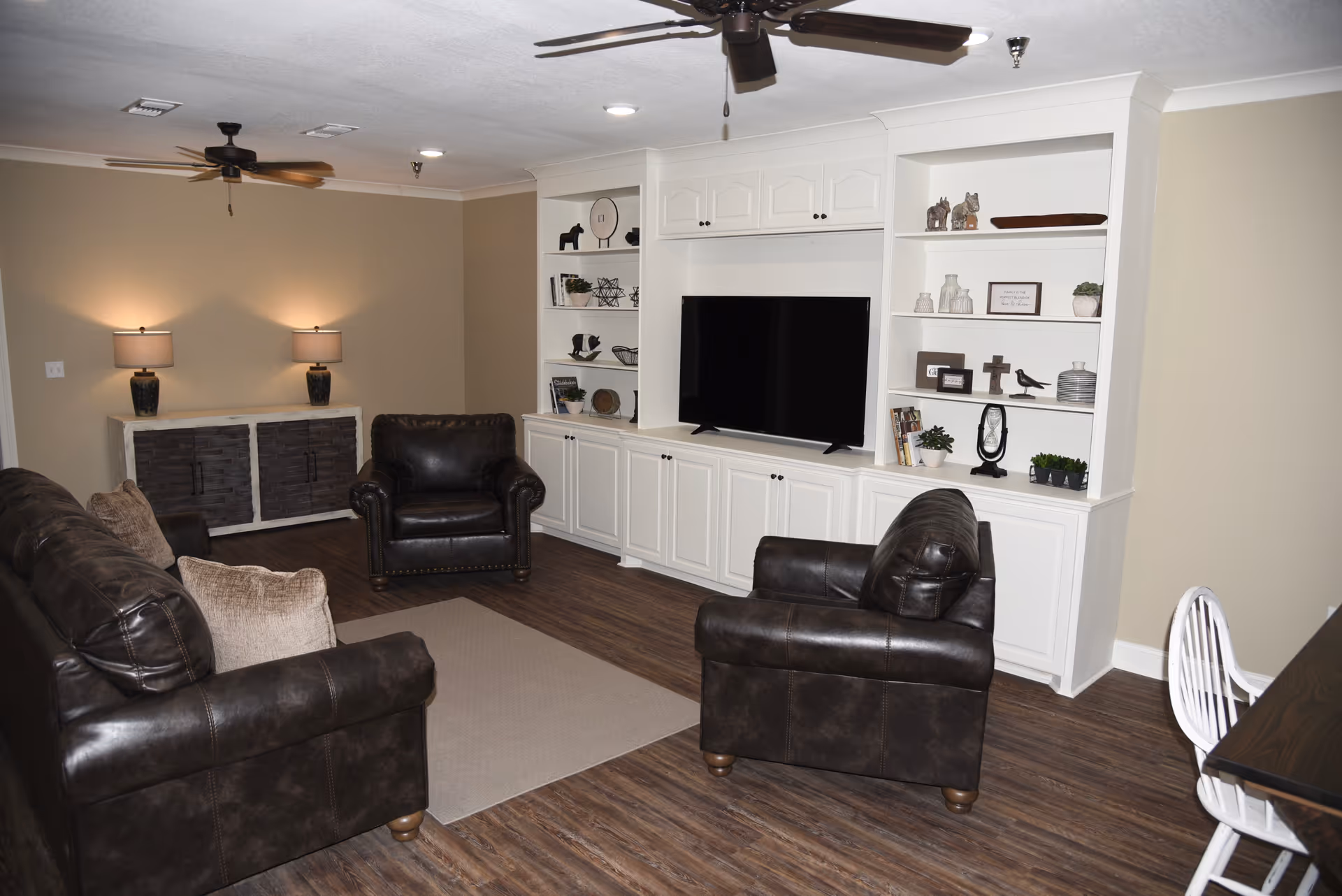 A cozy living room with dark brown leather sofas and armchairs arranged around a beige rug. The room features a built-in white entertainment center with shelves decorated with various ornaments and a flat-screen TV. Two table lamps with beige shades sit on a sideboard against a beige wall. The floor is wood with a dark finish, and ceiling fans with lights are mounted on the white ceiling.