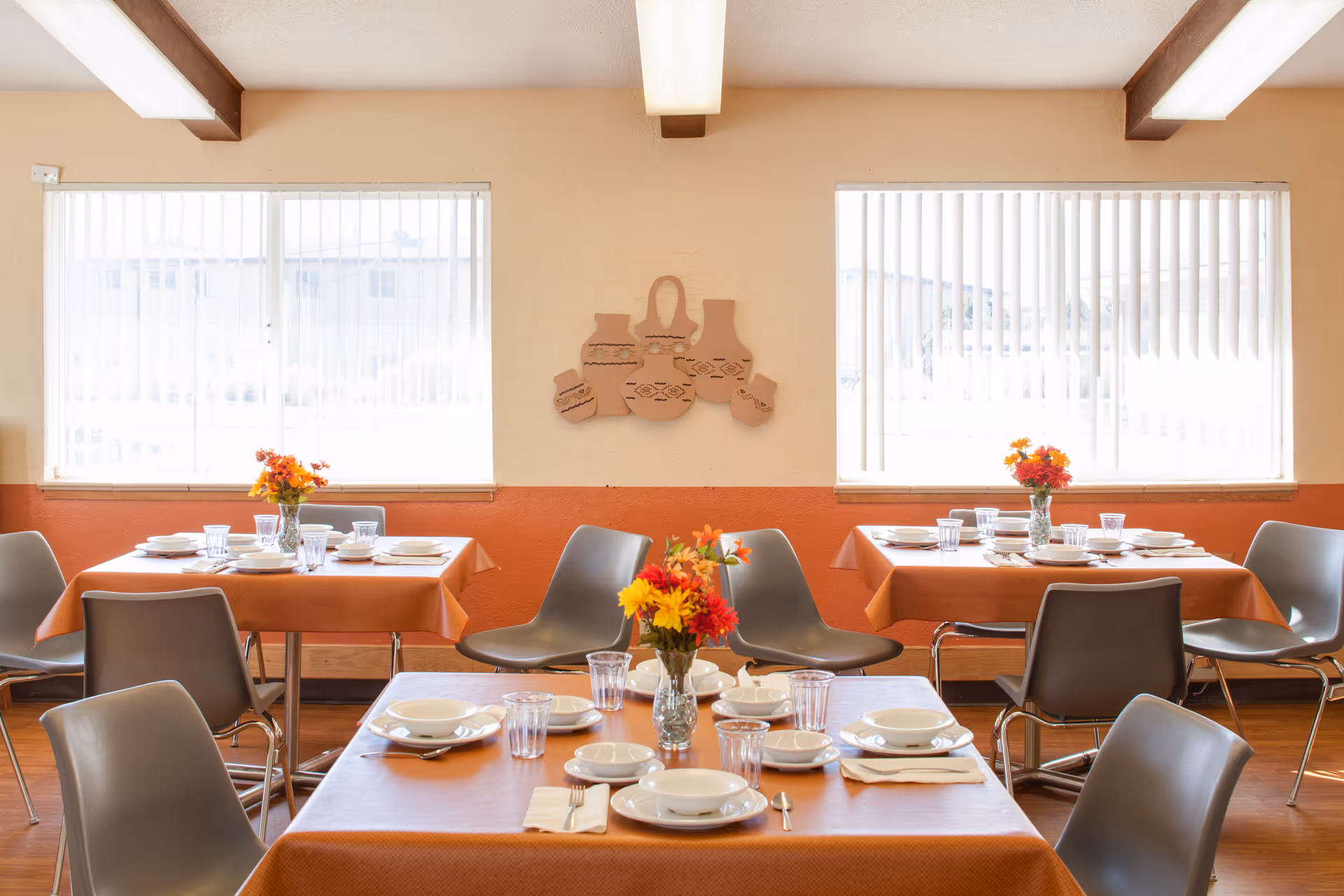 Dining room with orange-tablecloth tables set for meals, gray chairs, and flower centerpieces beneath large windows.