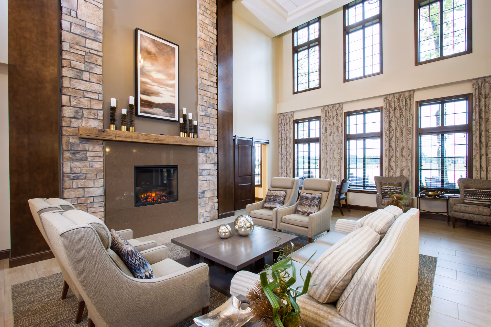 A spacious and well-lit living room with high ceilings and large windows. The room features a stone fireplace with candles and a framed picture above it. There are several comfortable armchairs and sofas arranged around a central coffee table with decorative spheres. The windows have patterned curtains, and there are additional chairs and tables near the windows.