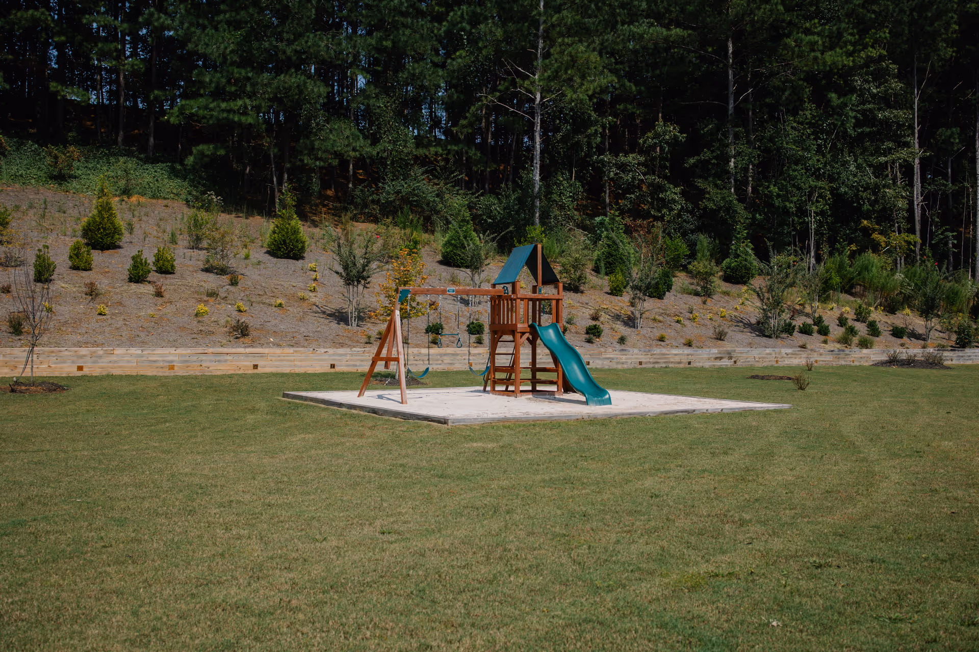 A wooden children's playset with a green slide and swings sits on a grassy field in front of a wooded hillside.