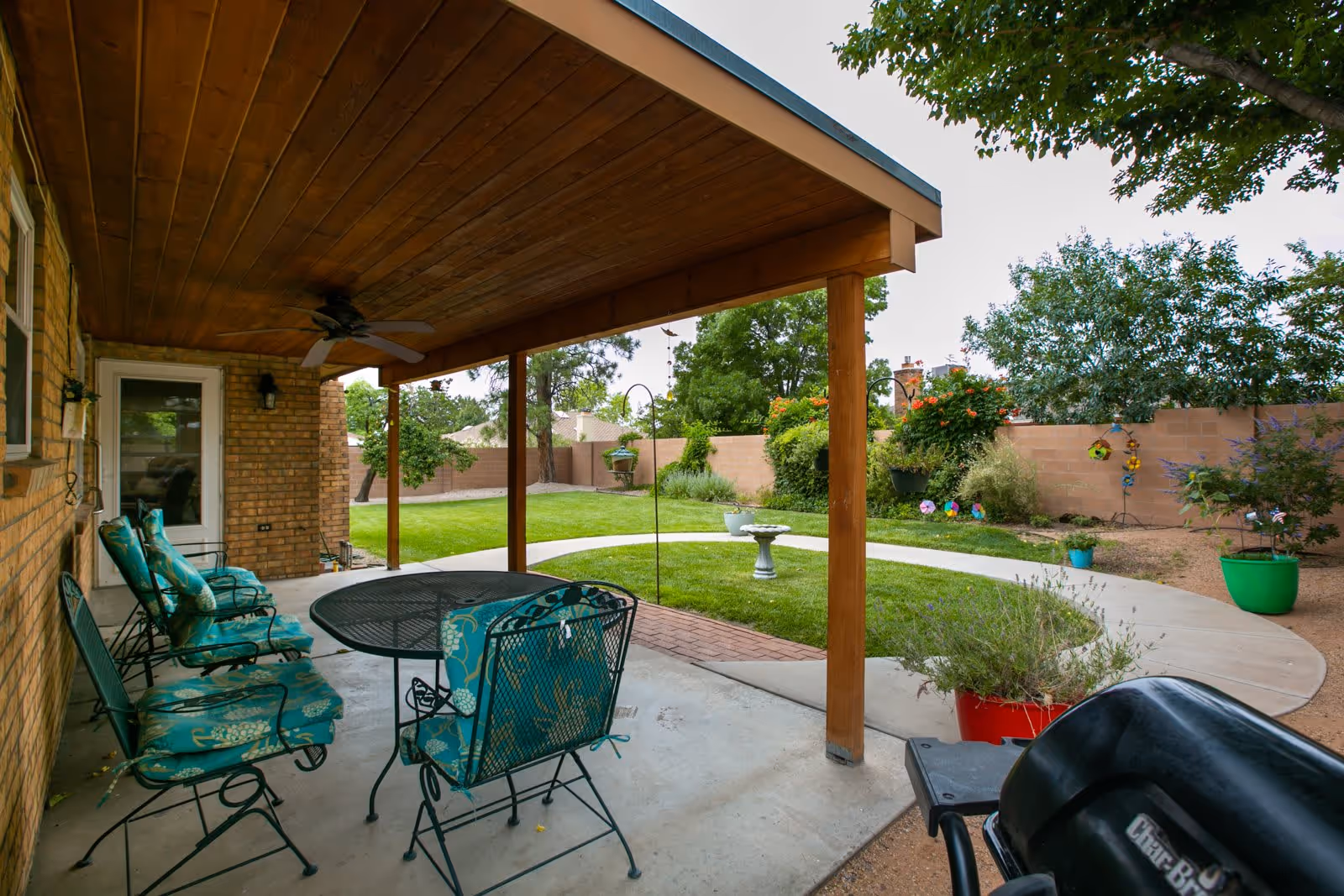 Covered patio area with a wooden ceiling and ceiling fan, featuring a round metal table and several cushioned metal chairs. The patio overlooks a well-maintained garden with a curved concrete pathway, green grass, various plants, and a birdbath. A black grill is visible in the foreground on the right side.