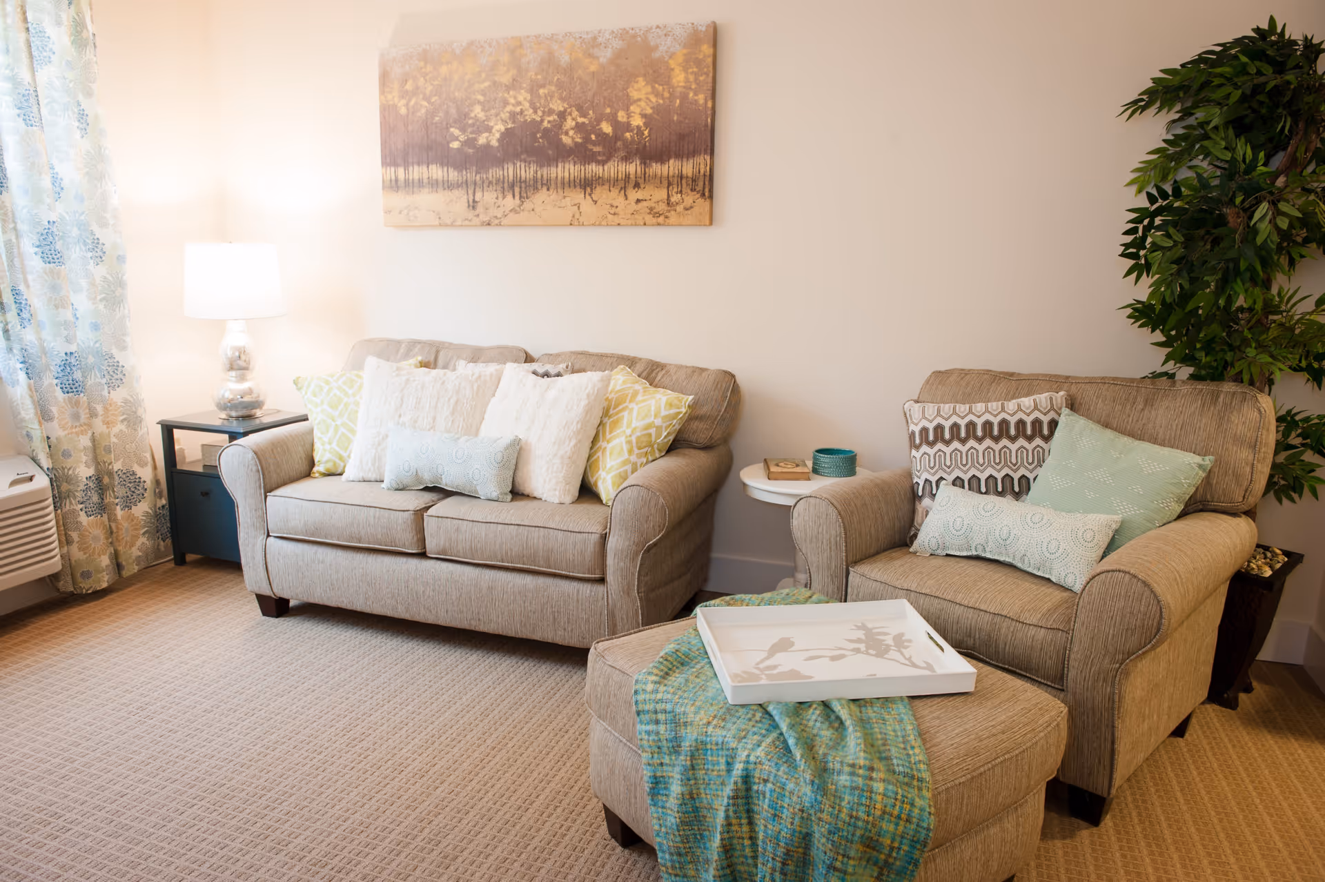 A cozy living room with a beige loveseat and armchair, both adorned with multiple decorative pillows. A matching ottoman with a blue-green throw blanket and a white tray sits in front of the armchair. To the left, there is a side table with a lamp and floral curtains on the window. A large potted plant is placed to the right, and a framed artwork of a forest scene hangs on the wall above the loveseat.