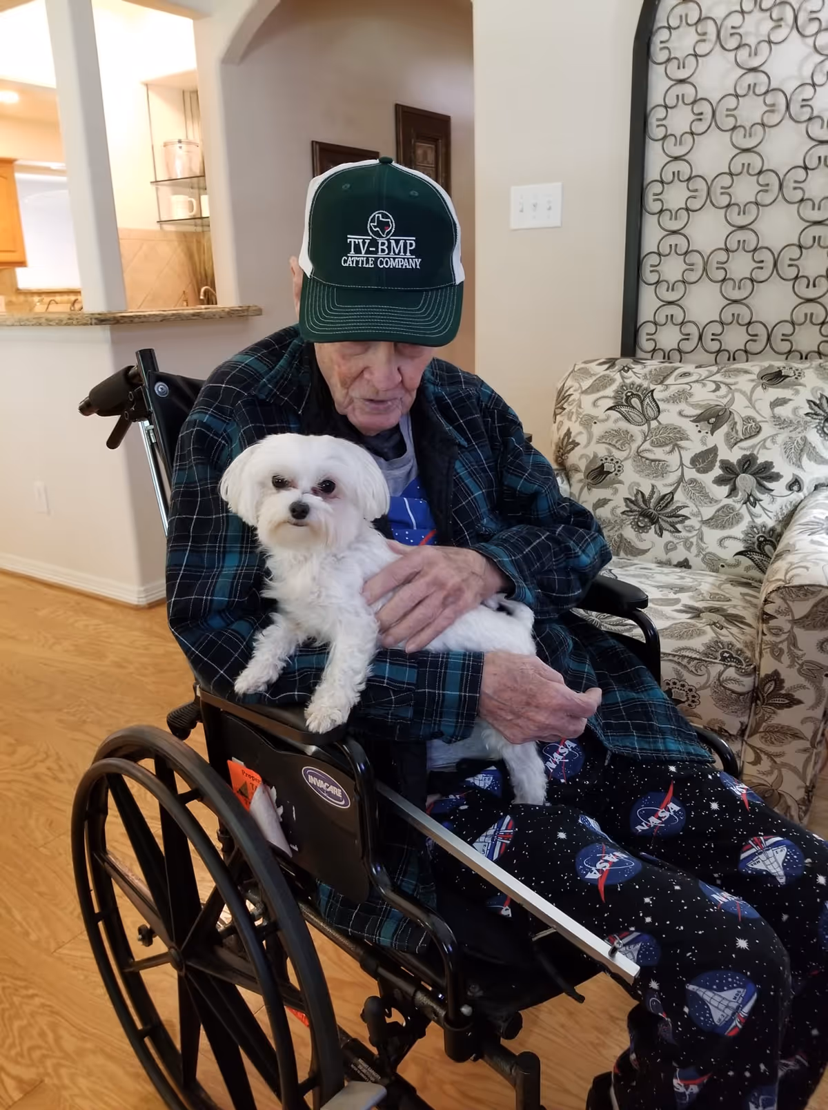 An elderly man sitting in a wheelchair indoors, wearing a green and white cap, a plaid shirt, and NASA-themed pants, holding a small white dog on his lap. The background shows part of a living room with a patterned couch and decorative wall art.