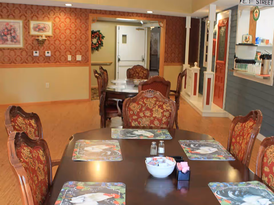 Dining area with a dark wooden table set with six ornate chairs featuring red and gold upholstery. The table has placemats with animal designs, a bowl with condiments, salt and pepper shakers, and a container with sugar packets. The room has wood flooring, patterned wallpaper on the upper half of the walls, and framed floral artwork. In the background, there is a doorway with a wreath and a counter area with coffee dispensers and cups.