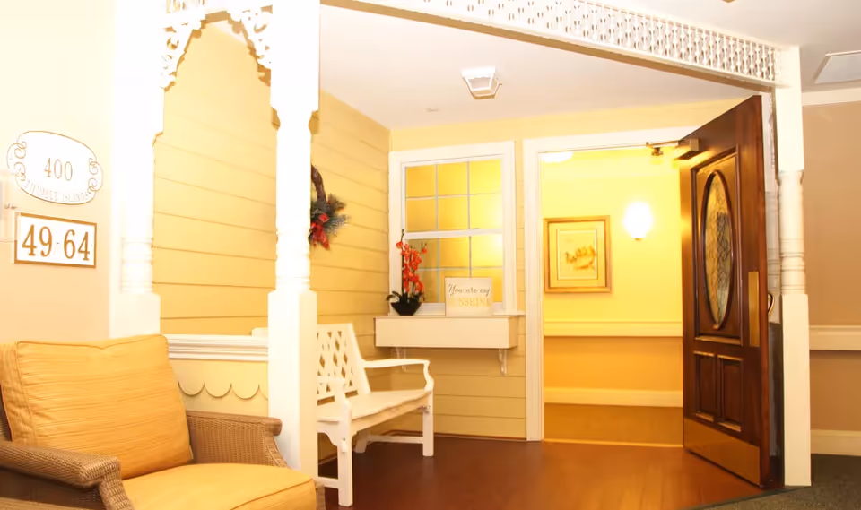 A cozy interior corner of a senior living facility with a cushioned armchair on the left, a white wooden bench against a yellow paneled wall, a small window with a flower pot and a sign that reads 'You are my sunshine', and an open wooden door leading to a warmly lit hallway with framed artwork on the wall.