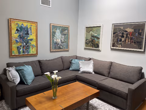 A cozy living room corner with a dark gray L-shaped sectional sofa adorned with light blue and white pillows. A wooden coffee table with a glass vase holding white calla lilies sits in front of the sofa. The walls are light gray and decorated with four colorful abstract paintings in gold and silver frames.
