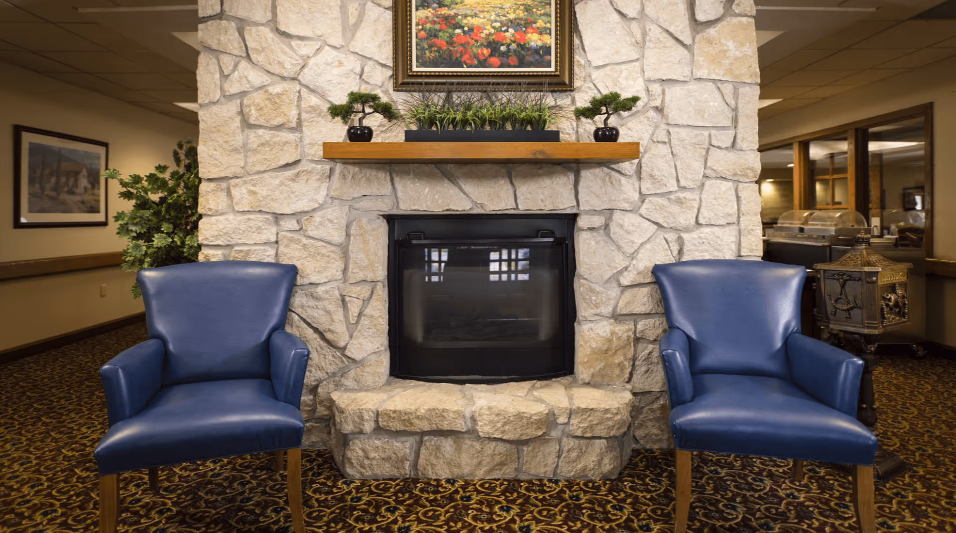 Two blue armchairs flank a stone fireplace with a wooden mantel and framed painting above in a common area.