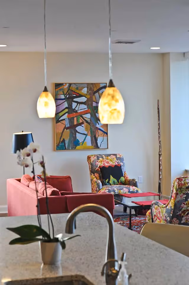 Kitchen island and sink in the foreground with pendant lights over a living area featuring a red sofa, floral armchairs, and abstract wall art.