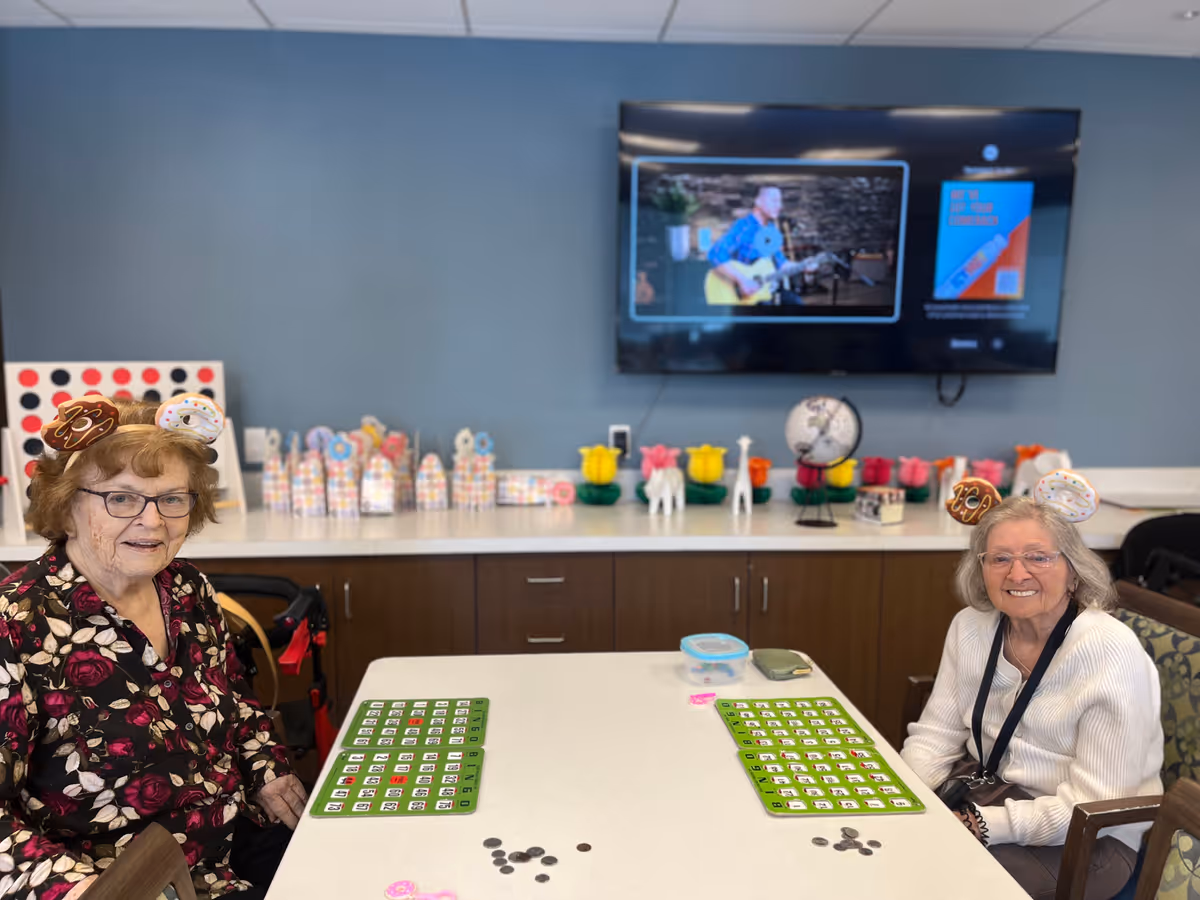 Two elderly women sitting at a table playing bingo in a recreational room. Both women are wearing festive headbands with donut designs. Behind them is a counter with colorful decorations and a large flat-screen TV mounted on a blue wall displaying a man playing guitar.