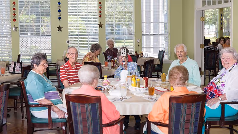 A group of elderly people sitting around a round dining table in a bright room with large windows. The table is set with drinks, condiments, and napkins. The room has light-colored walls decorated with hanging star ornaments and numbered table signs. The people appear to be engaged in conversation and enjoying their time together.