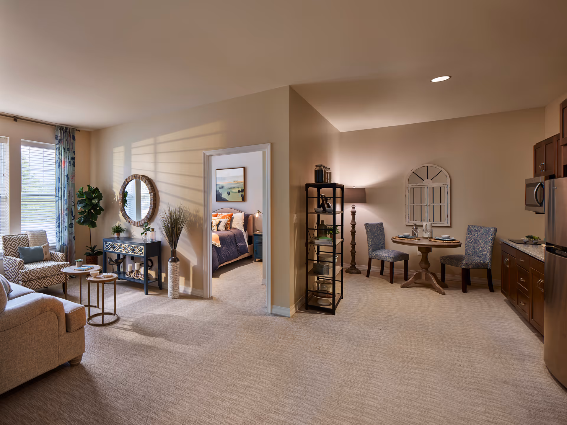 A cozy living space with a sitting area featuring a beige sofa, patterned armchair, and small round tables near large windows with blue floral curtains. A decorative console table with a round mirror and plants is against the wall. To the right, there is a small dining area with a round wooden table, two upholstered chairs, a floor lamp, and a shelving unit. A kitchen area with wooden cabinets and stainless steel appliances is visible on the far right. A doorway leads to a bedroom with a bed, nightstand, and artwork on the wall.
