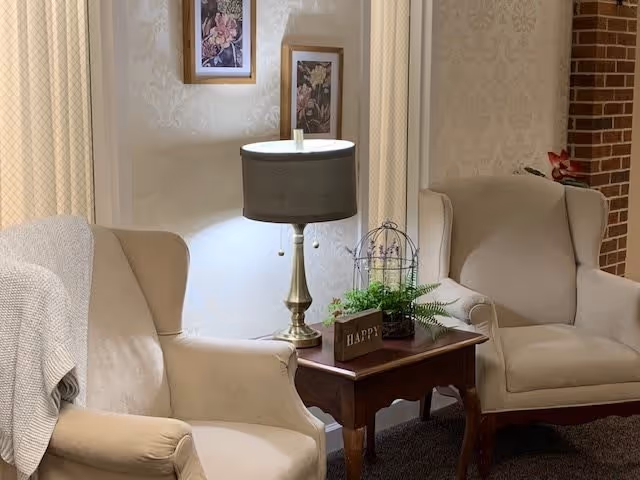 A cozy seating area with two beige upholstered armchairs separated by a wooden side table. The table holds a brass lamp with a dark lampshade, a small decorative birdcage, a green plant, and a wooden block sign that says 'HAPPY'. The wall behind features light patterned wallpaper and two framed floral pictures. A light-colored blanket is draped over one armchair.