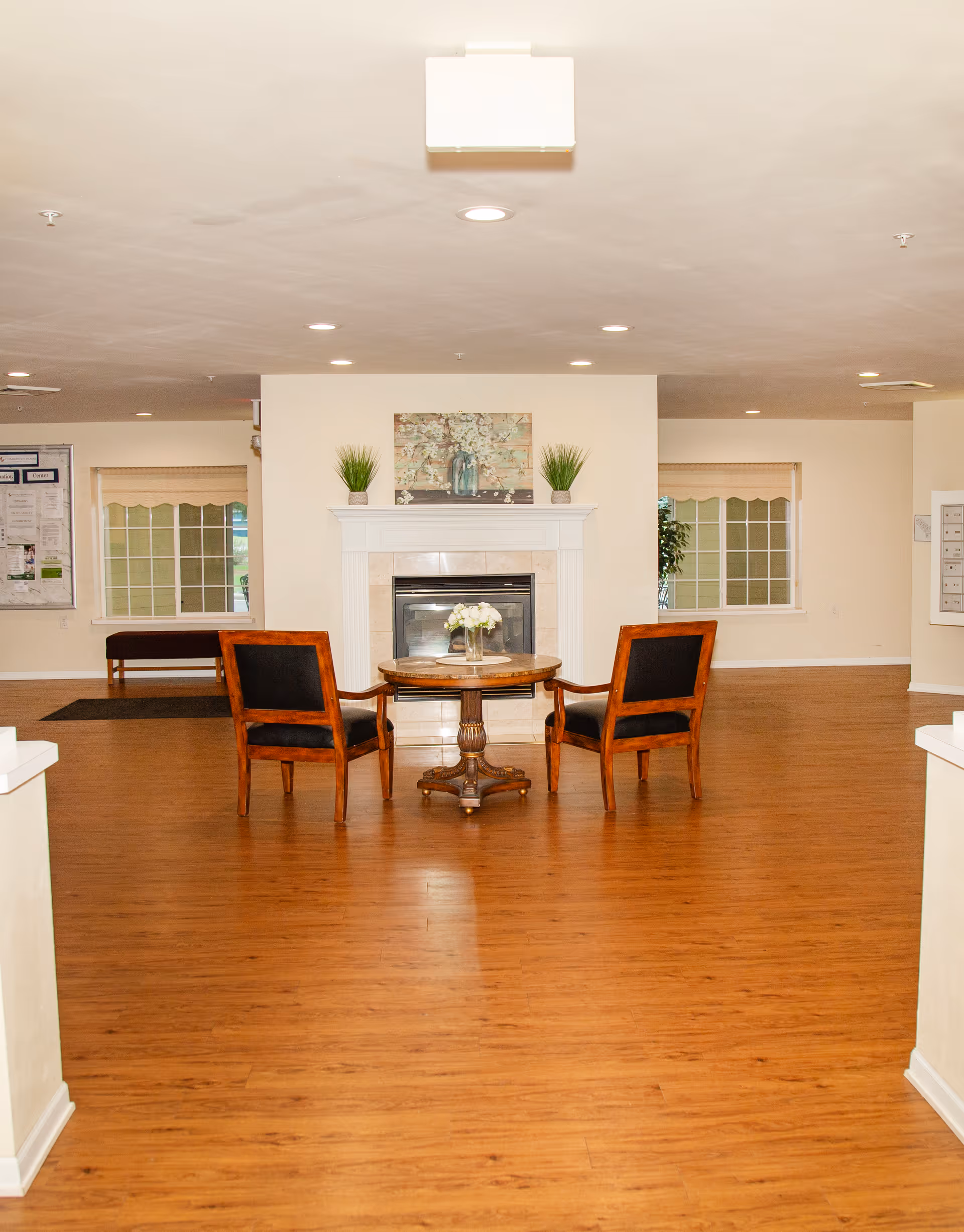 A spacious room with wooden flooring featuring a small round table with a flower vase on top and two wooden chairs with black cushions facing a white fireplace. The fireplace mantel has two potted plants and a decorative painting above it. There are windows with beige blinds on either side of the fireplace and a bulletin board on the left wall.