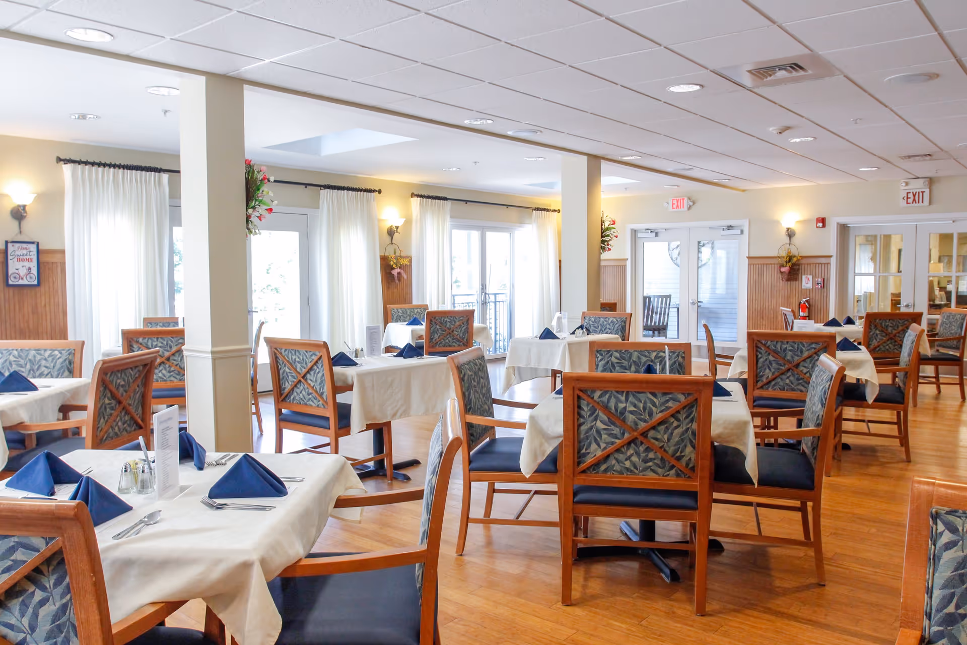 A bright and spacious dining room with multiple tables covered in white tablecloths, each set with blue folded napkins, silverware, and menus. The room features wooden chairs with patterned upholstery, large windows with white curtains allowing natural light to fill the space, and light-colored walls with wall sconces and floral decorations.