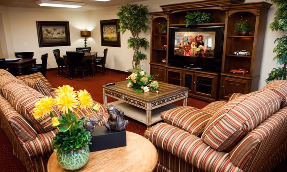 A cozy living room area with two striped sofas facing a wooden entertainment center with a television. The room has a red carpet, a wooden coffee table with a floral arrangement, and a round side table with a green vase holding yellow flowers. In the background, there are two framed paintings on the wall, a lamp, a tall plant, and a dining table with chairs.