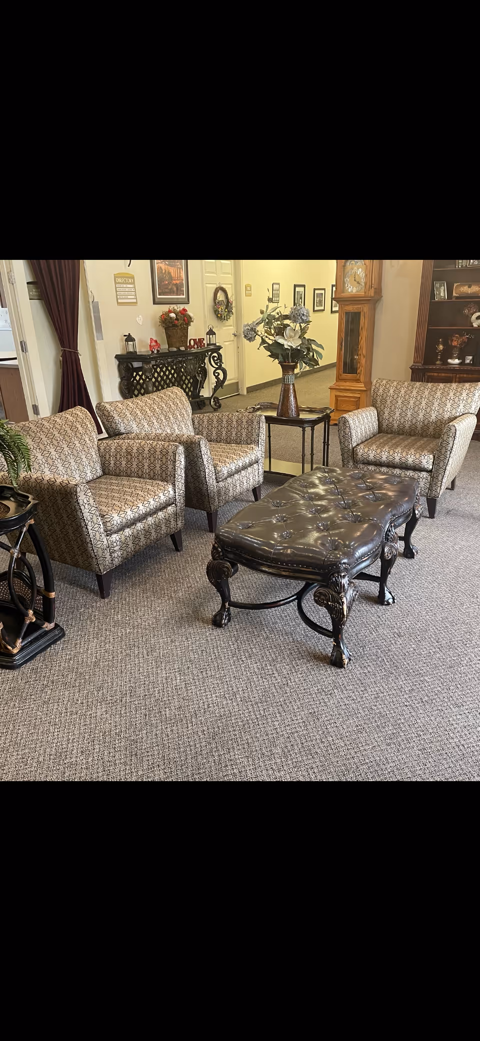 A cozy sitting area in an assisted living facility featuring three patterned armchairs arranged around a dark, ornate leather-tufted coffee table. The room has beige carpeting and is decorated with a tall grandfather clock, a side table with a vase of flowers, and a decorative console table with various ornaments and framed pictures on the wall.