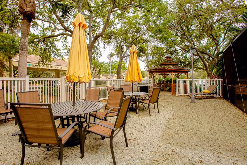 Outdoor seating area with round tables and chairs under yellow umbrellas, surrounded by trees and a white fence. A wooden gazebo is visible in the background along with a lounge chair and a lamp post.