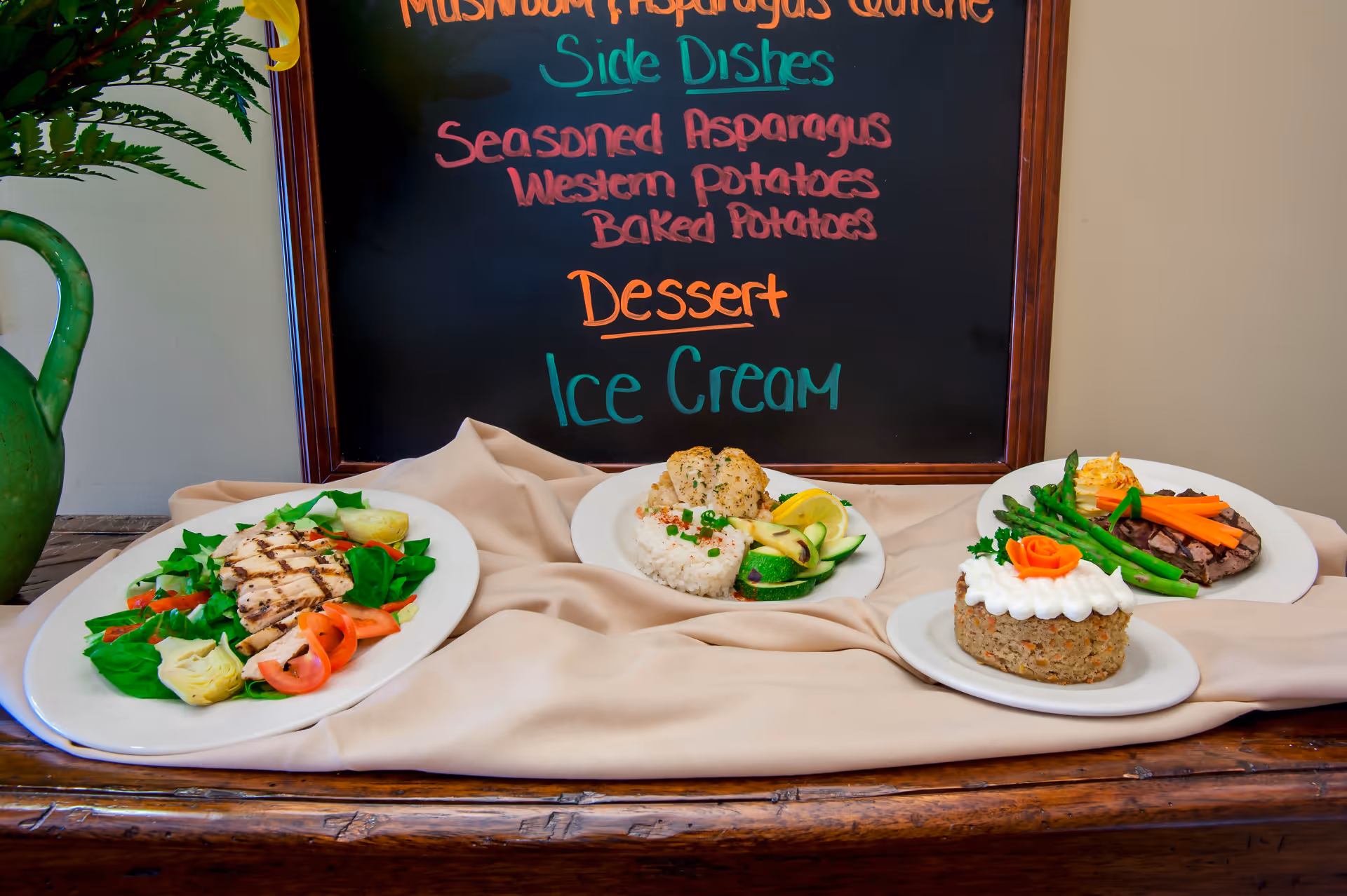 A display of plated meals on a wooden table with a beige cloth, featuring grilled chicken salad, a dish with rice and vegetables, a meatloaf with white frosting and a carrot garnish, and a steak with asparagus and carrot sticks. Behind the plates is a chalkboard menu listing side dishes and dessert options including seasoned asparagus, western potatoes, baked potatoes, and ice cream.
