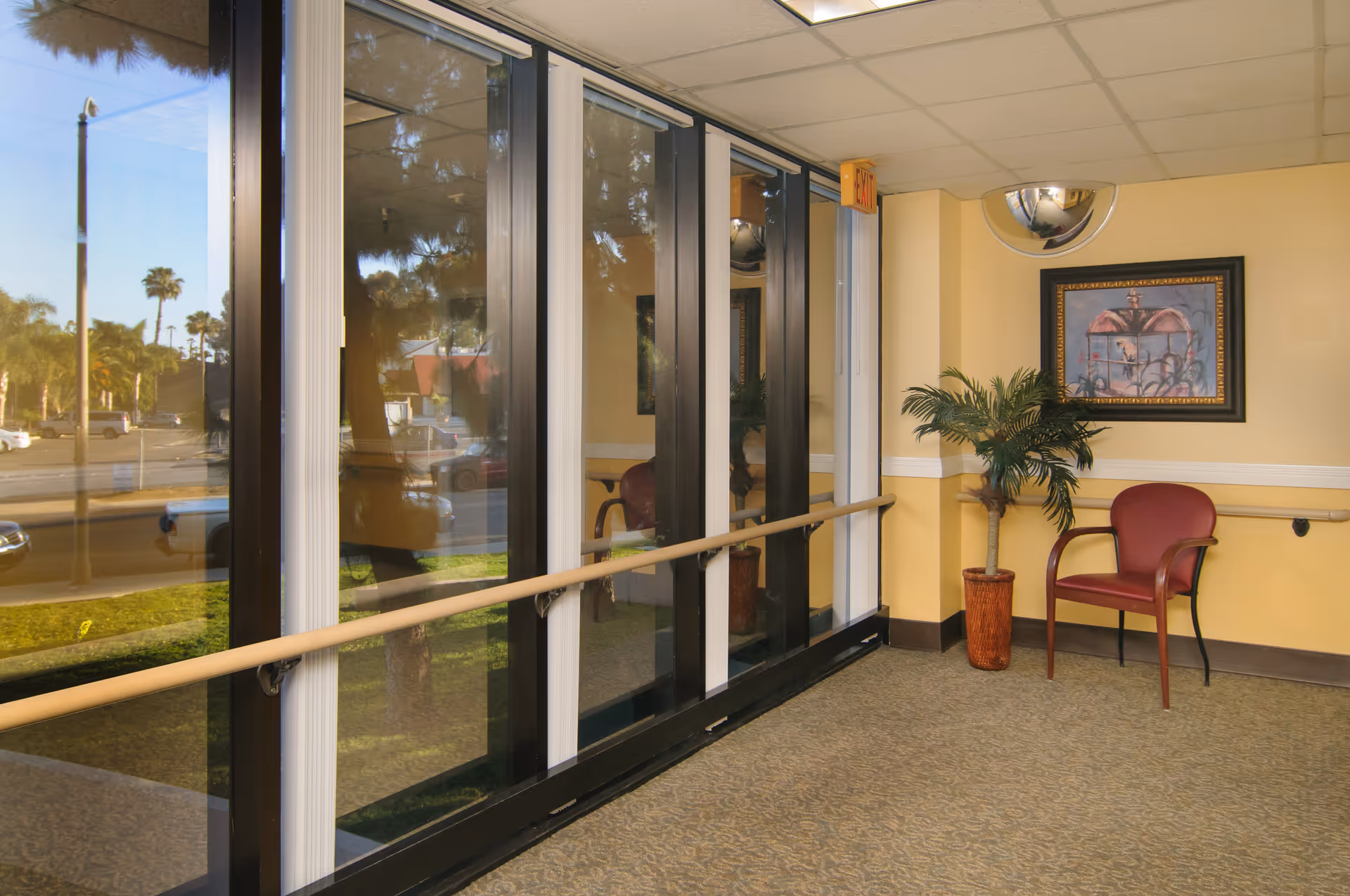 Interior hallway with large floor-to-ceiling windows on the left side, a handrail running along the wall, a red chair, a potted plant, and a framed painting on the right wall. The hallway has beige walls and carpeted floor.