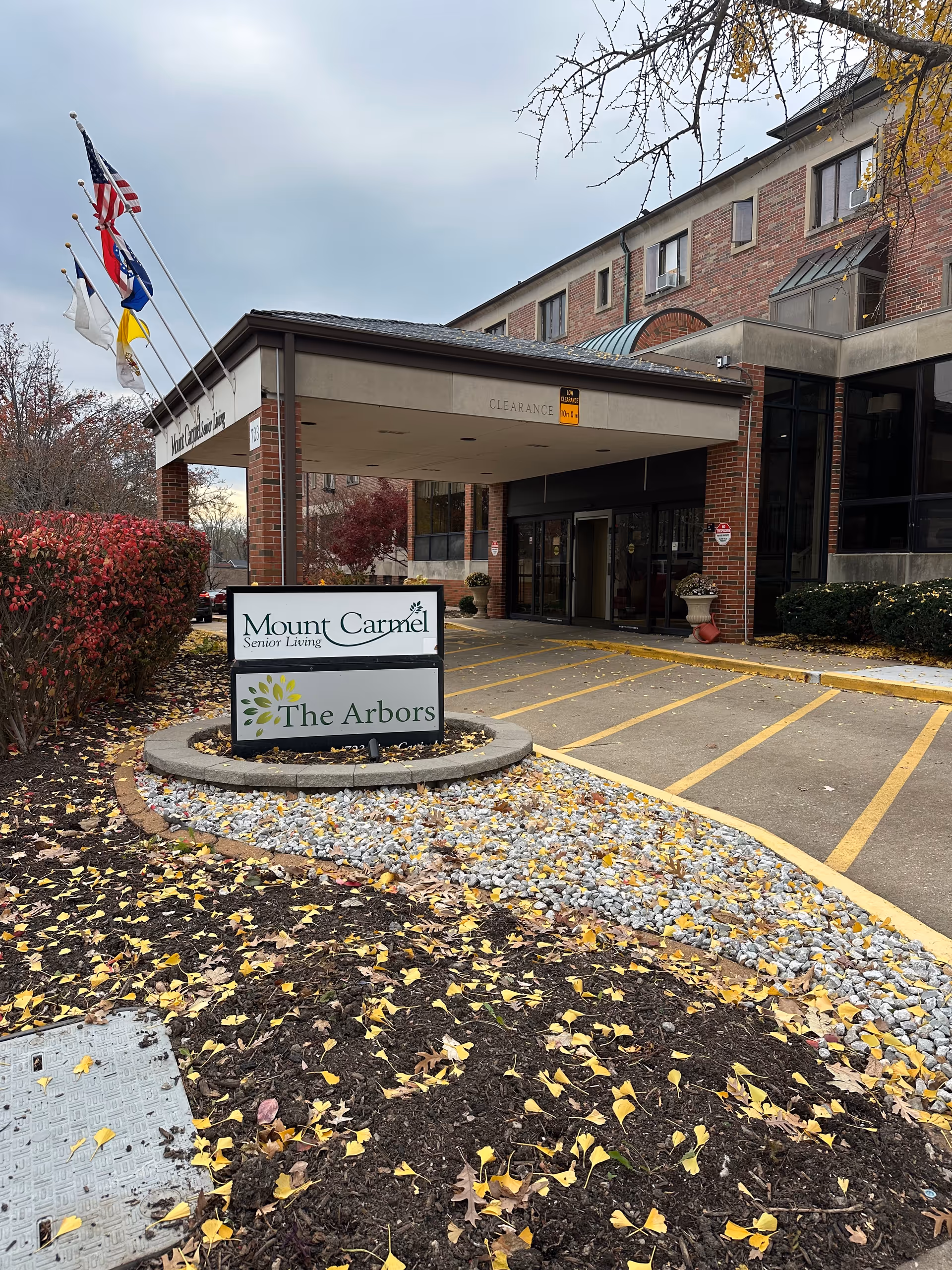 Exterior view of the entrance to Mount Carmel Senior Living facility with a covered drop-off area. There is a sign in front that reads 'Mount Carmel Senior Living' and 'The Arbors'. Several flags are flying on poles to the left of the entrance. The ground is covered with fallen yellow leaves and landscaping includes bushes and rocks.