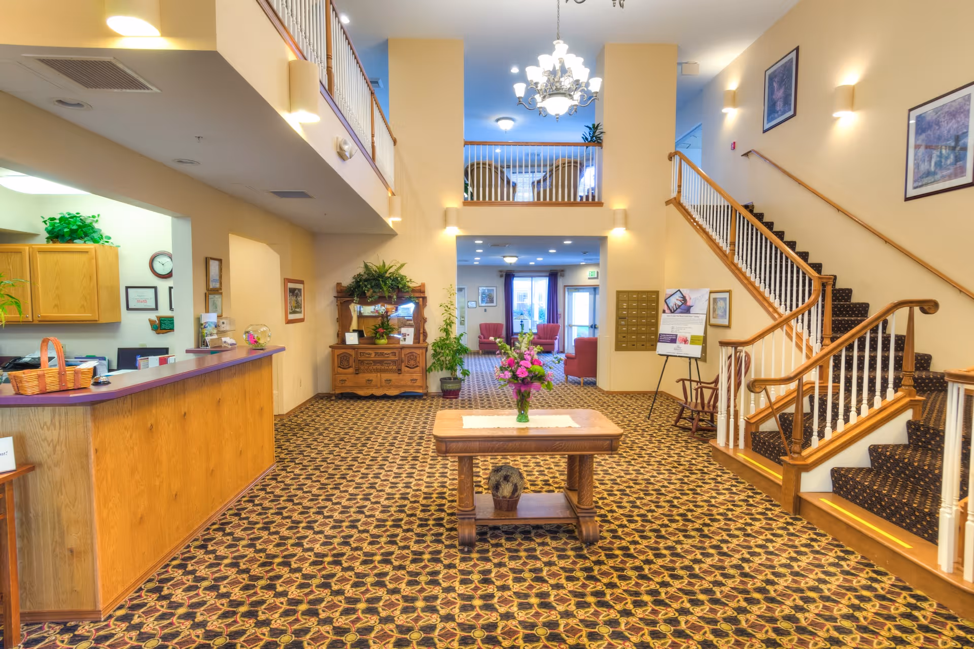 Interior view of a senior living facility lobby with a patterned carpet, a wooden reception desk on the left, a wooden table with a flower vase in the center, a staircase with wooden handrails on the right, and seating areas visible in the background under bright lighting.